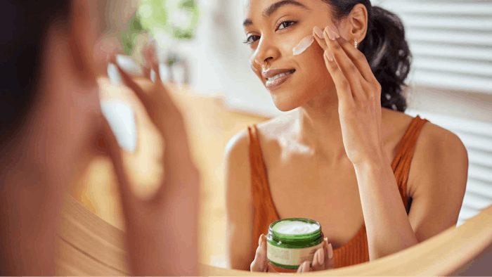 medium-toned woman applying natural skin cream in mirror smiling