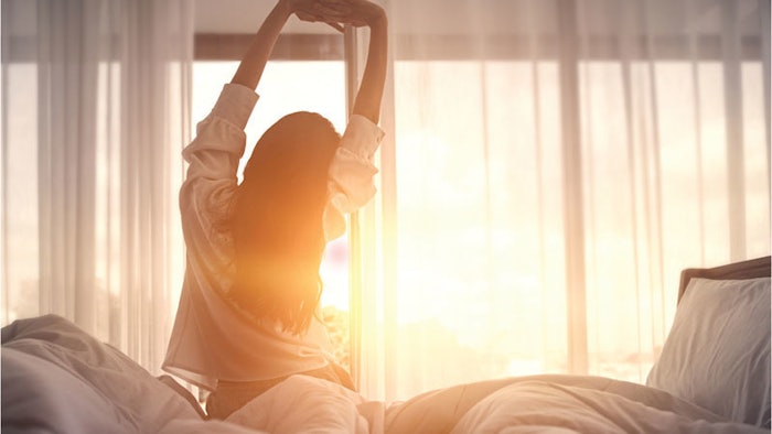 Woman sitting and stretching on bed in front of a window showing the sun rising.