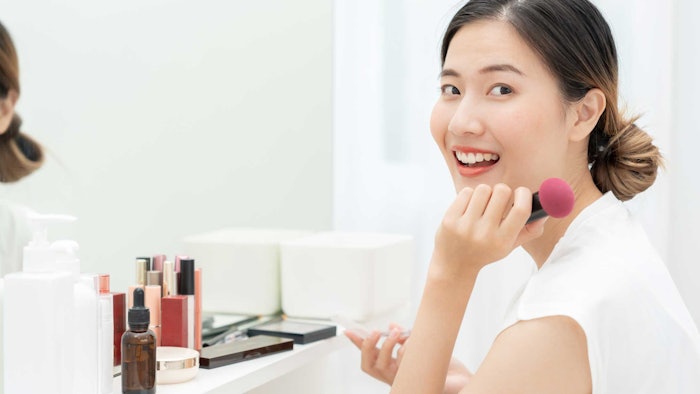 happy Asian woman seated at makeup mirror cosmetics on shelf smiling at camera