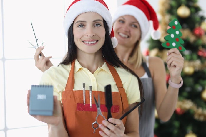 Two hairstylists behind a Christmas tree wearing Santa hats and holding a mini-calendar, shears, combs and a Christmas tree ornament.
