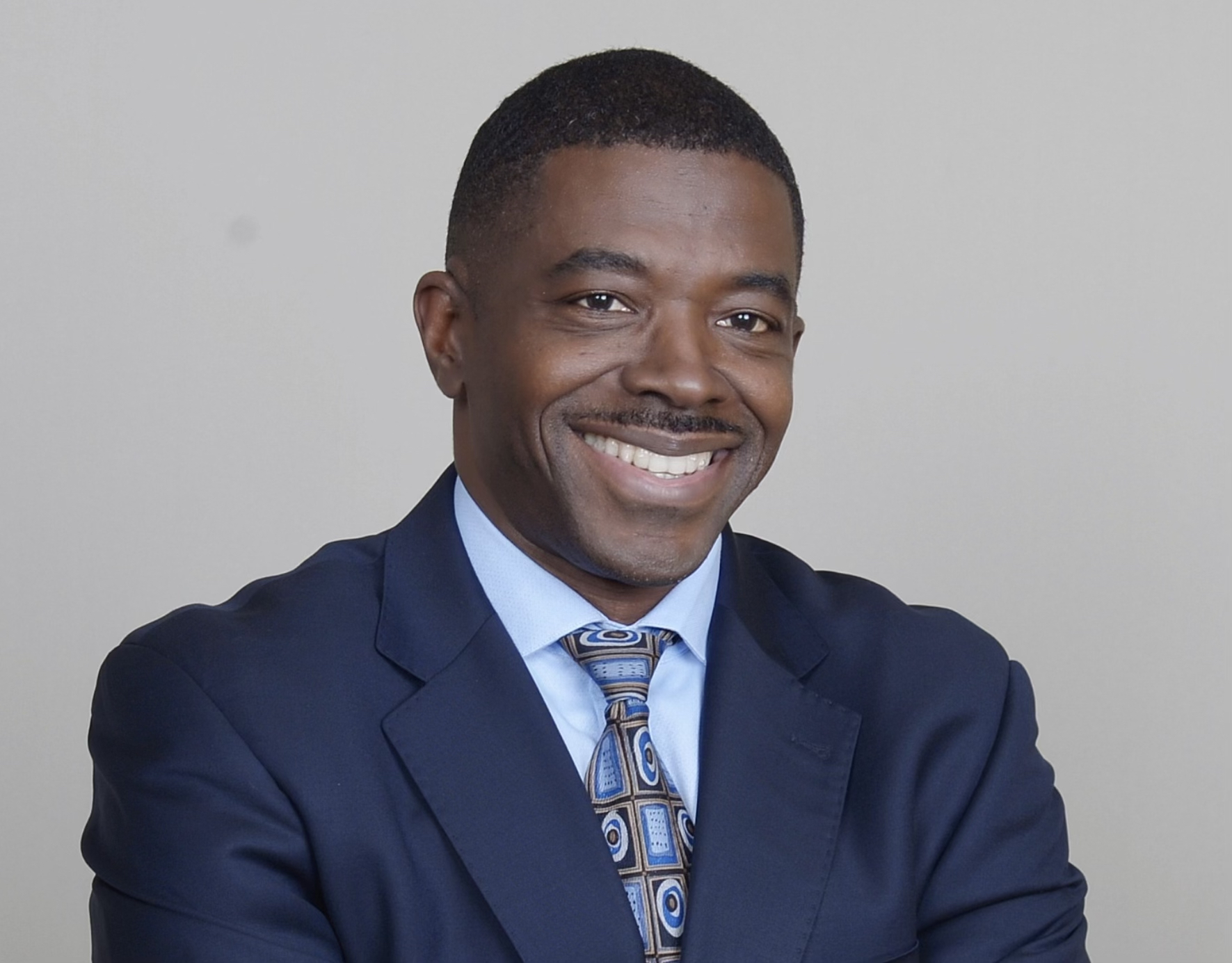 Professional smiling man in navy suit and patterned tie against neutral background
