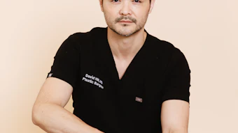 Professional headshot of male plastic surgeon in black medical scrubs against neutral beige background
