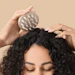 Person using a metal scalp massager on curly black hair against a neutral beige background