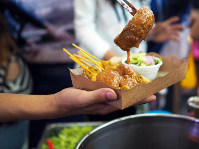 Hand holding paper container of grilled meat skewers with golden fries and dipping sauce at outdoor food stall
