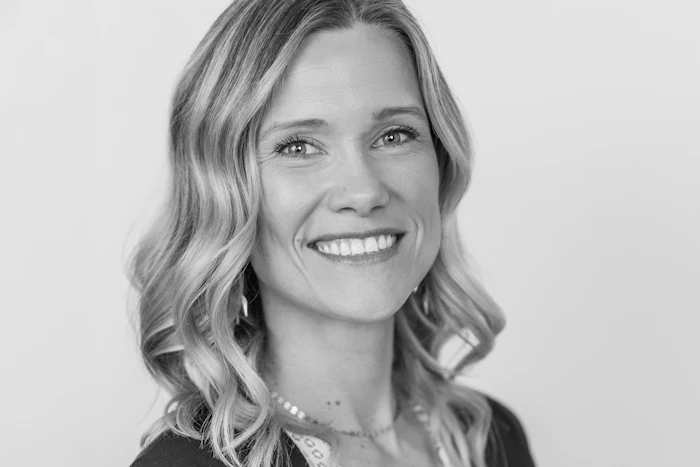 Professional headshot of smiling woman with wavy blonde hair wearing dark top and pearl necklace