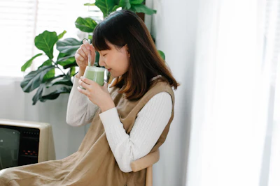 Woman in beige vest and white shirt sipping tea from green mug near potted plant