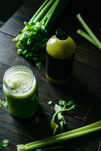 Fresh celery stalks and homemade green celery juice in a glass jar and dark bottle on wooden surface