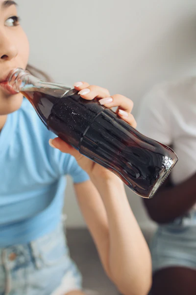 Young person in blue shirt drinking dark cola from glass bottle