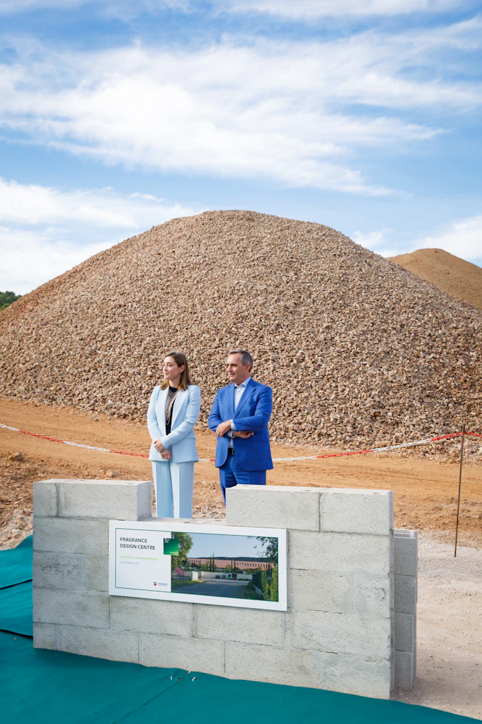 Two professionals stand at construction site with large gravel pile and Fragrance Design Centre signage