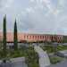 Modern terracotta building with cypress trees, landscaped grounds, and parking area under cloudy sky