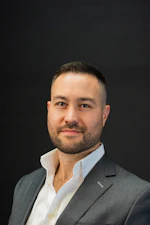 Professional headshot of a man in gray blazer and white shirt against dark background