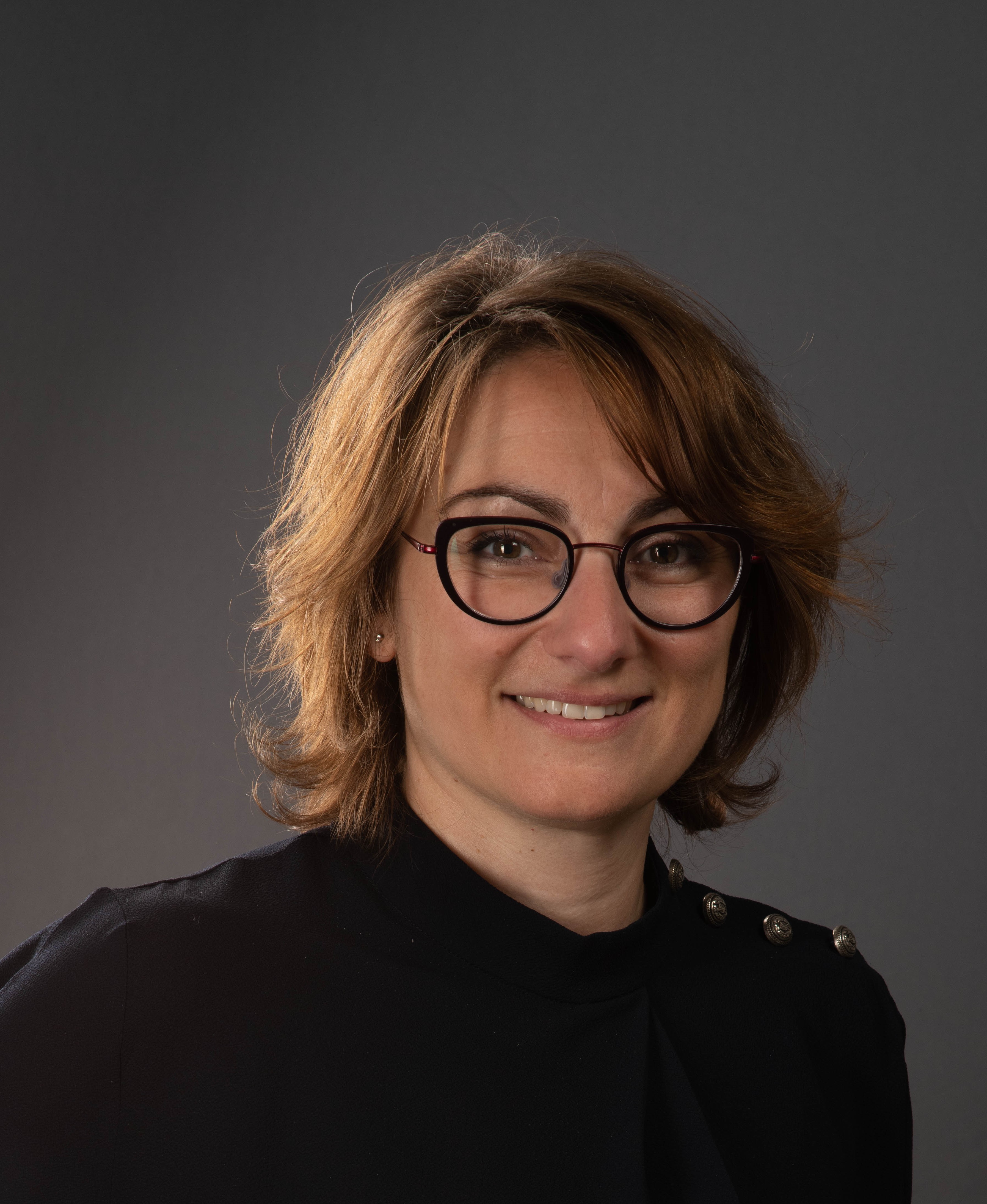 Professional headshot of a smiling woman wearing glasses and a black blazer against a gray background