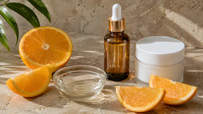 Skincare products with amber dropper bottle and white jar displayed with fresh orange slices and green leaves on textured surface