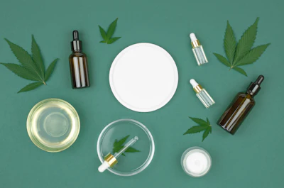 Cannabis-infused skincare products including dropper bottles, serums, and creams arranged with fresh cannabis leaves on teal background