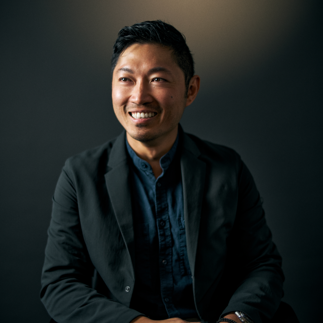 Professional headshot of an Asian man smiling at camera, wearing black blazer and dark blue shirt against dark background
