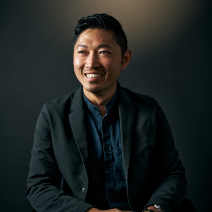 Professional headshot of an Asian man smiling at camera, wearing black blazer and dark blue shirt against dark background