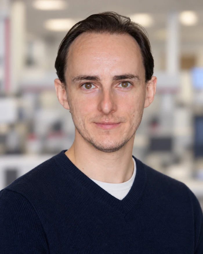 Professional headshot of a man with dark hair wearing a navy sweater in an office setting