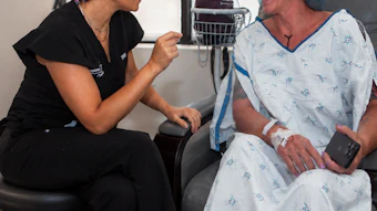 Healthcare provider in scrubs with patterned head covering speaking with smiling patient in hospital gown during medical consultation