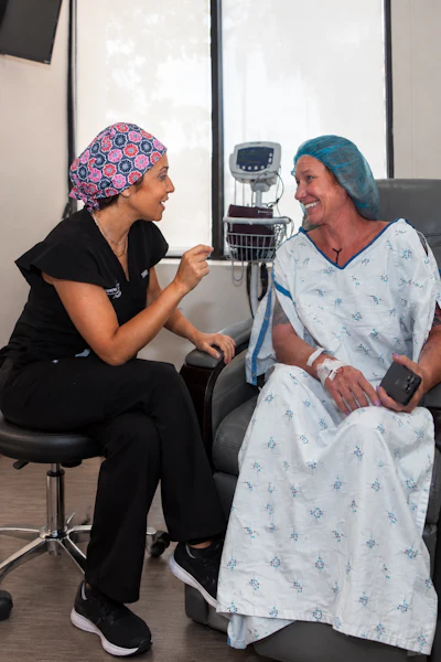 Healthcare provider in scrubs with patterned head covering speaking with smiling patient in hospital gown during medical consultation