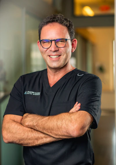Male healthcare professional wearing glasses and black medical scrubs with arms crossed, smiling in clinical setting