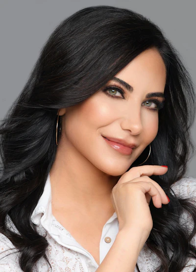 Professional portrait of a woman with long black wavy hair, wearing a white shirt and gold hoop earrings, smiling against a gray background