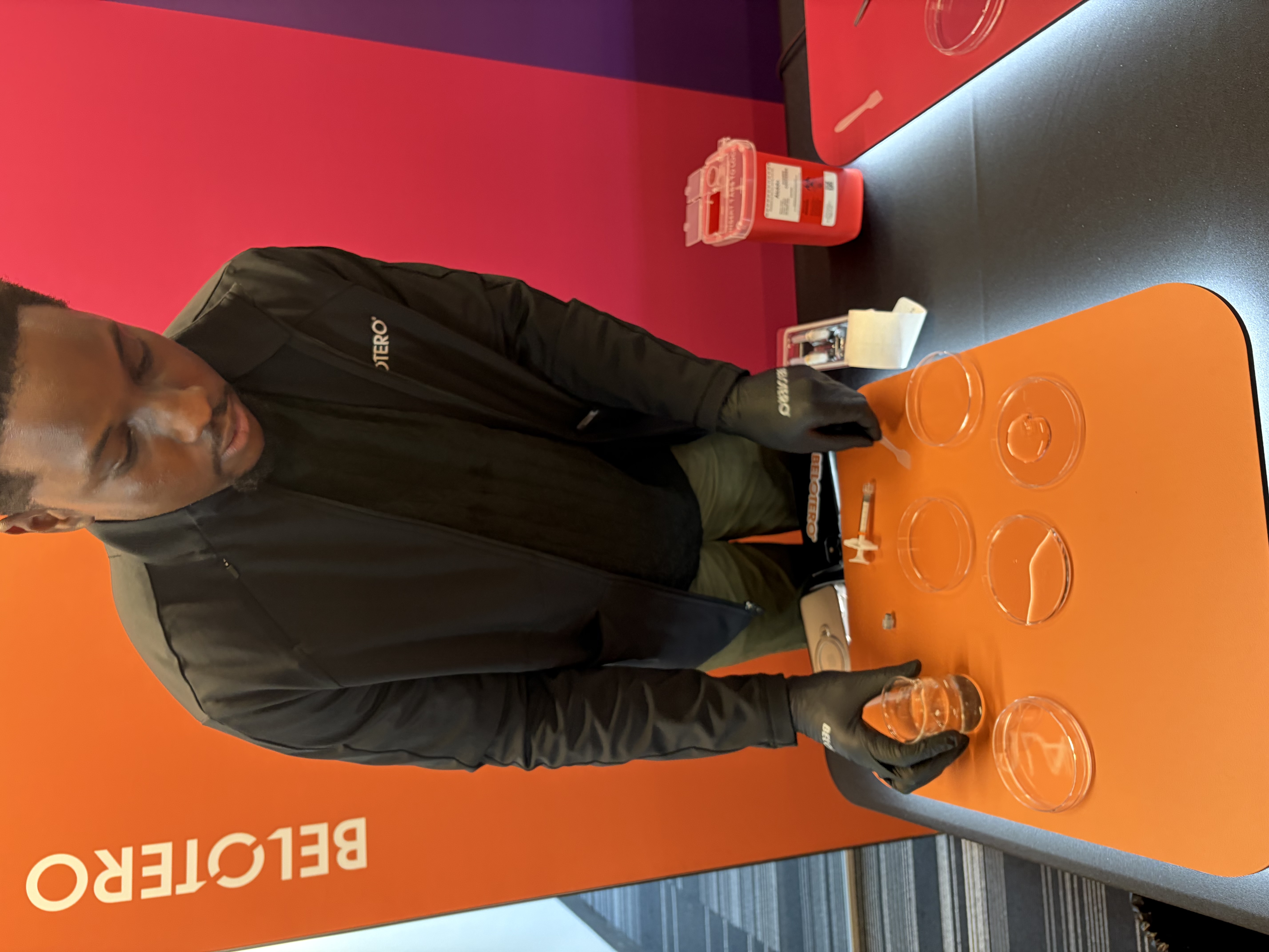 Man in black jacket demonstrating laboratory procedure with petri dishes at Belotero branded event table