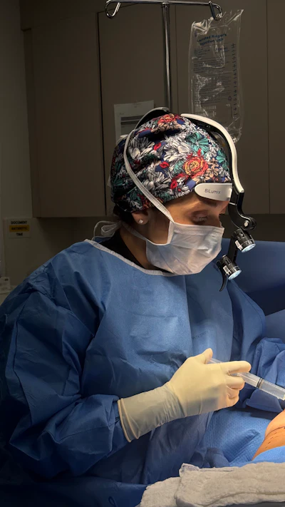Healthcare professional wearing blue scrubs, white mask, and floral surgical headband with BiLumix headlight during medical procedure