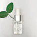 Clear serum bottle with silver dropper cap next to fresh green leaves on white background