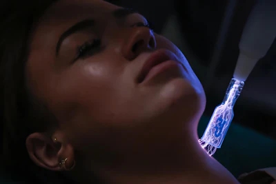 Person receiving blue light facial treatment with glowing handheld device against dark background