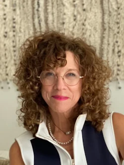 Professional woman with curly brown hair wearing glasses and a navy and white sleeveless vest, seated on a white couch in a modern office setting
