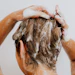 Person lathering hair with shampoo while washing, hands visible applying white foam to wet brown hair
