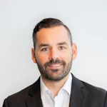 Professional headshot of a bearded man in black blazer and white shirt against neutral background